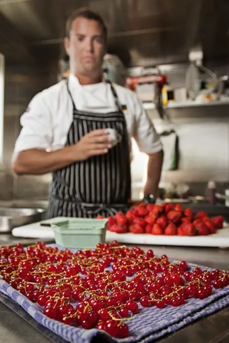 Pastry chef holding a cup of espresso while standing in kitchen with assorted berries being prepped on counter