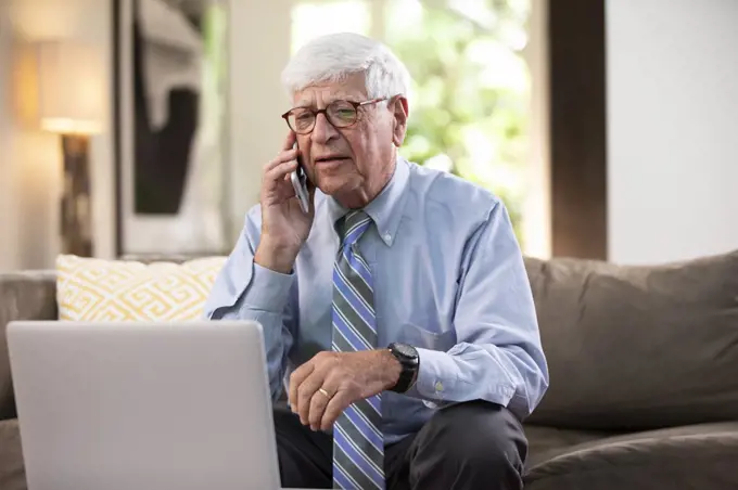 Mature Caucasian Male doctor practicing tele-medicine from his home, using cell phone and laptop computer, Talking to patient