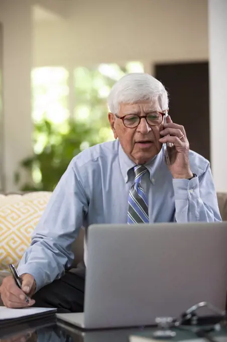 Mature Caucasian Male doctor practicing tele-medicine from his home using laptop computer, Talking to patient on cell phone taking notes