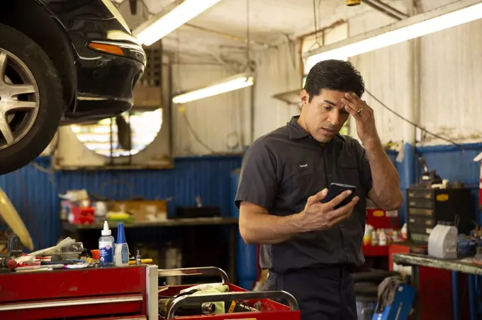 Hispanic car mechanic working in auto shop, having migraine headache, using mobile phone to check in with tele-medicine doctor 