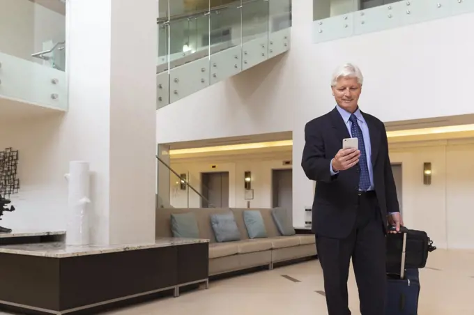 Mature Caucasian man looking at cell phone while walking through hotel lobby with luggage