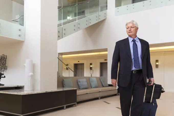 Mature Caucasian man holding cell phone walking through hotel lobby with luggage, looking off camera 