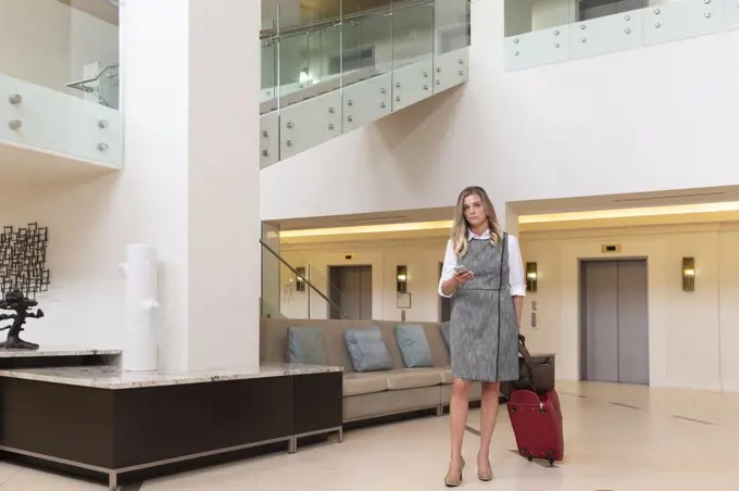 Young Caucasian woman with cell phone walking through hotel lobby with luggage, looking at camera