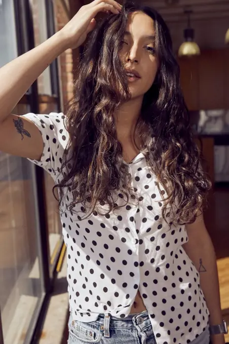 portrait of young ethnic woman wearing polka dot blouse and cut off jean shorts standing by bank of windows in downtown loft 