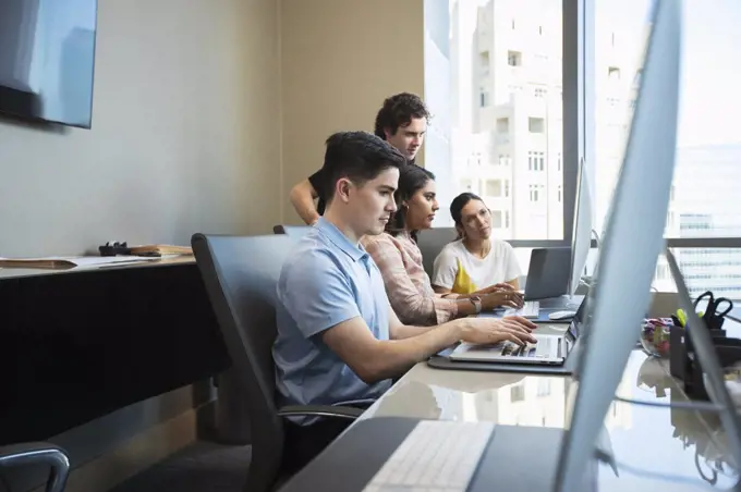 Young man using laptop computer in office conference room, co-workers having discussion in background 