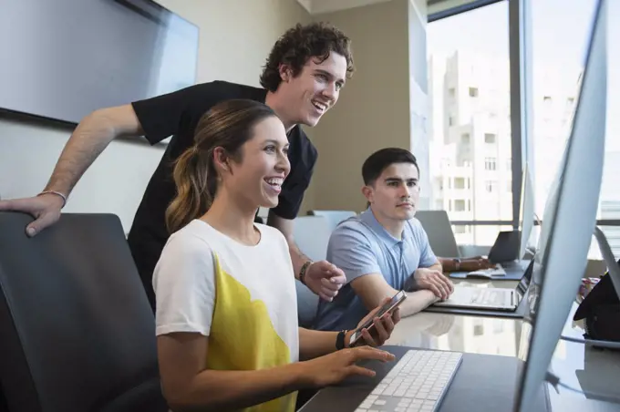 Young woman using computer in office holding mobile phone, showing male co-worker something on her screen with co-workers working in background 