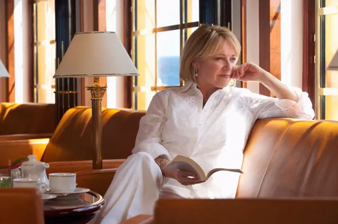 Mature woman reading book and relaxing on a leather couch in a ship lounge