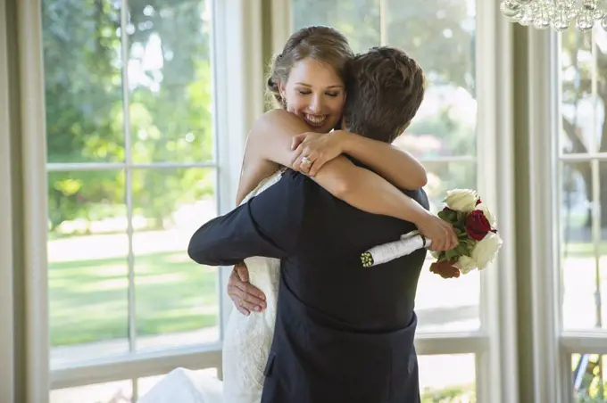 Bride and groom in an embrace, groom lifting up bride standing by large bank of windows 