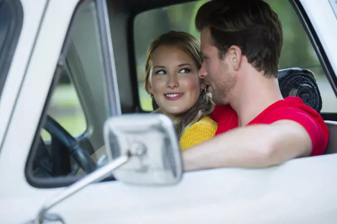 Young couple sitting in vintage pick up truck , guy with his arm around the girl while she looks over at him smiling. 