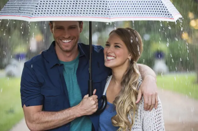 Young couple walking through park with umbrella in the rain, lovingly looking at one another  