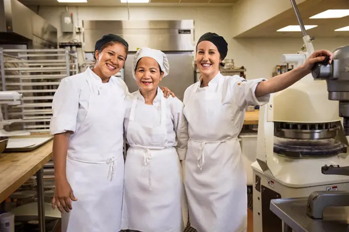 Portrait of three smiling female bakers in kitchen surrounded by culinary appliances. 