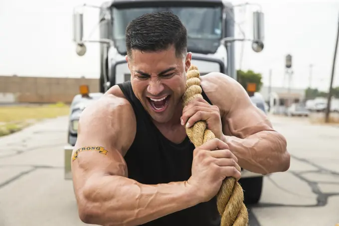 Muscular Hispanic man wearing tank top with Strong” tattoo on his bicep, Straining to pull semi truck with large tow rope 