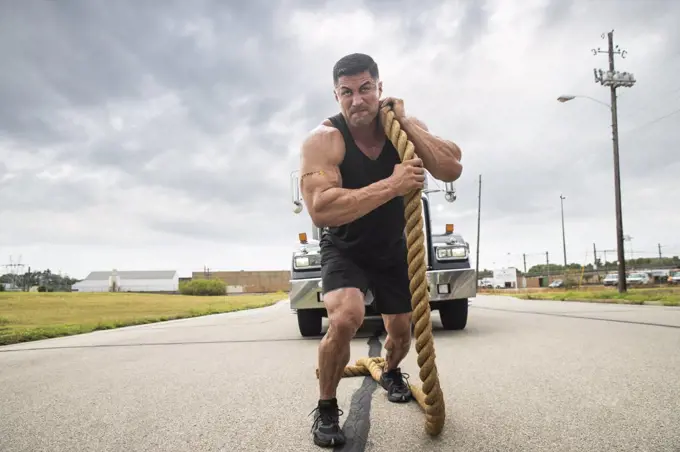 Muscular Hispanic man wearing tank top with Strong” tattoo on his bicep, Pulling semi truck with large tow rope 