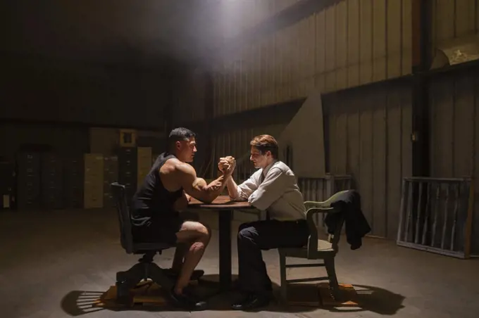 Two men preparing to have miss-matched arm wrestling contest in dingy warehouse 