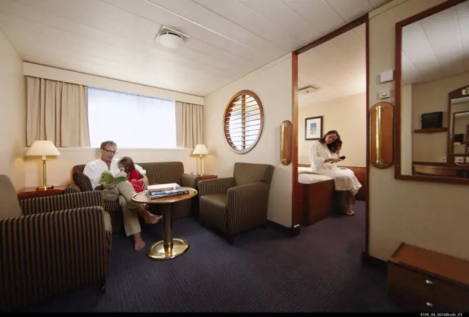 Family relaxing in cabin on eco luxury cruise ship, Father reading book to daughter while mother brushes hair in adjoining room in cabin of cruise ship