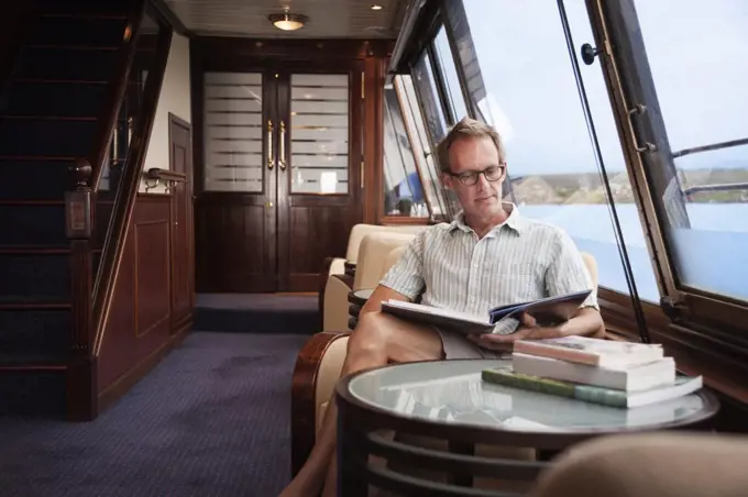 Man reading book while sitting in chair by window on cruise ship