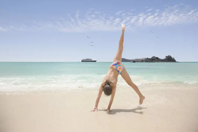 Teenage girl doing a cartwheel on the beach, cruise ship in ocean in background, Galapagos Islands