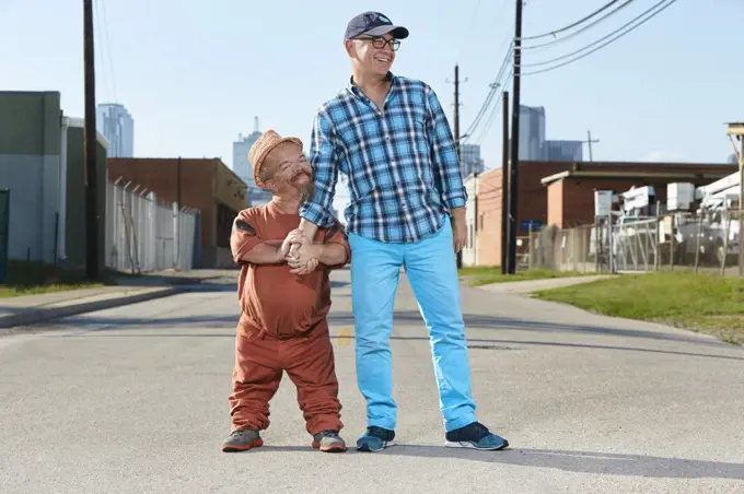 Portrait of male dwarf little person hugging his friend, standing in middle of street. 