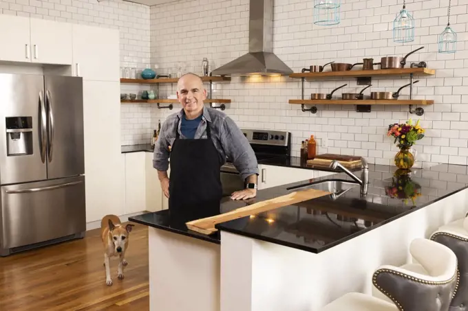 Portrait of man wearing apron in kitchen with his dog next to him, looking into camera with confidence. 