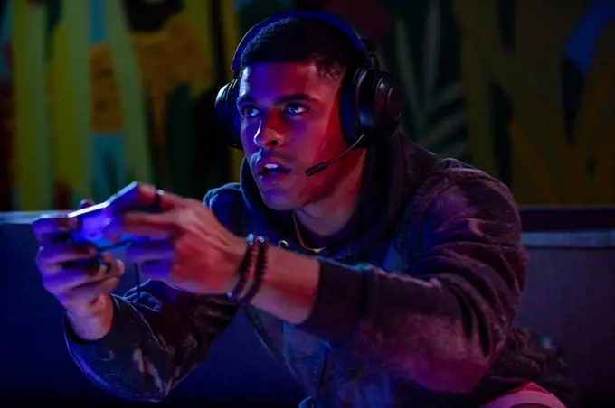 Front view of man with short hair holding a game controller and headset lit with blue light on a colorful background.