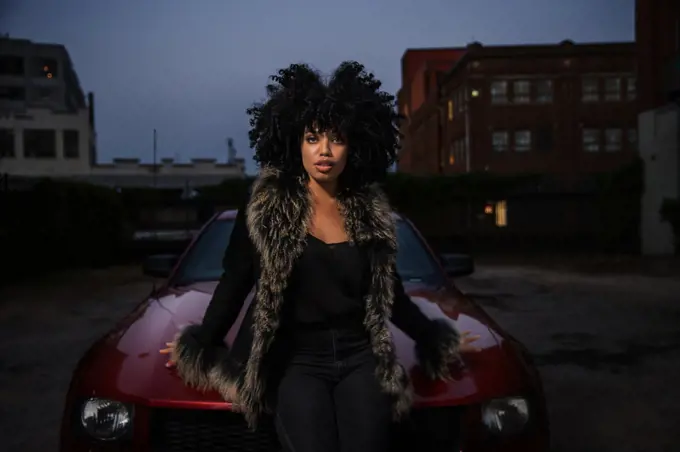 Confident young African American woman sitting on the hood of her car looking into camera.