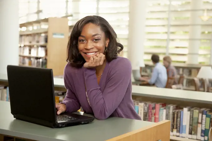 Smiling female student standing at bookshelf in library with laptop, looking at camera. 