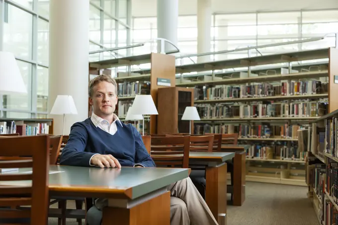 Portrait of college professor sitting at table in library looking at camera with confidence 