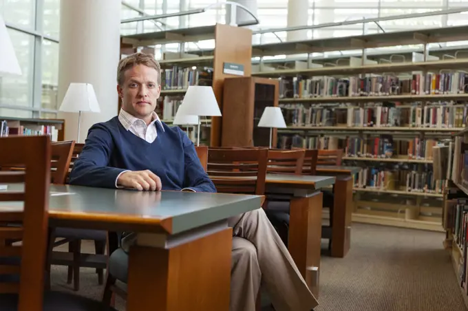 Confident high school teacher sitting in library looking at camera