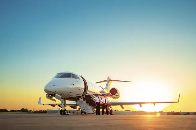 Two Pilots standing next to Jet on tarmac of small airport  at sunset