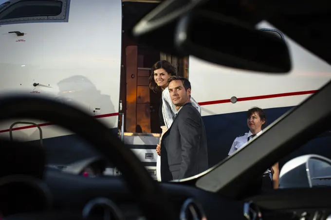 Successful couple boarding private Jet looking back over shoulders on tarmac of airport, view from drivers seat of sports car  parked next to plane on tarmac 