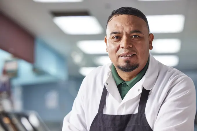 Smiling portrait of butcher wearing hair net behind counter of meat market of grocery store 