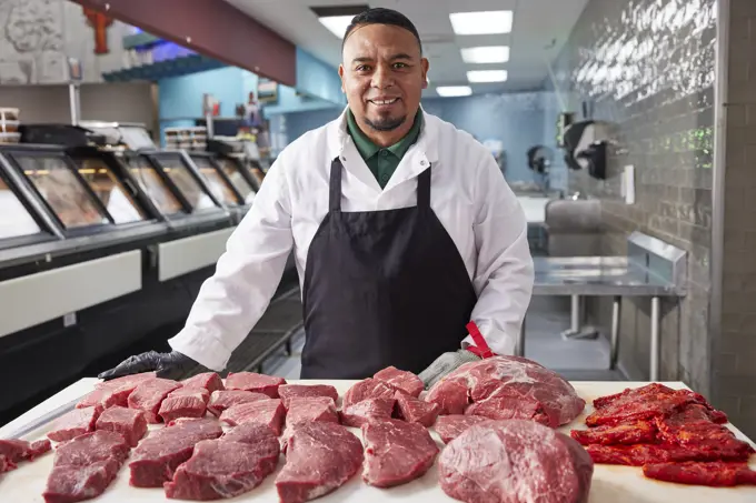 Smiling portrait of butcher wearing hair net behind counter of meat market of grocery store with selection of cuts of steak 