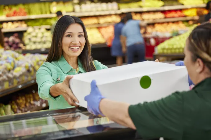 Portrait of smiling female customer with baker handing her customer custom cake order from bakery counter in grocery store, customers in produce section in background 