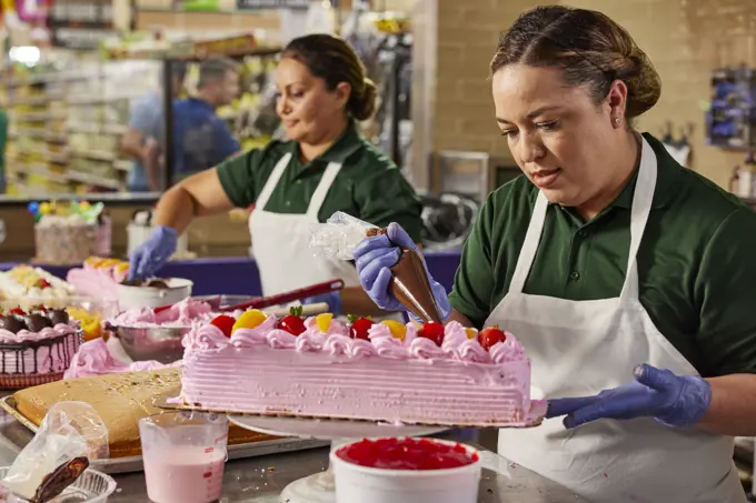 Female cake decorators putting finishing touches on layer cake in latin market bakery, piping on chocolate icing 