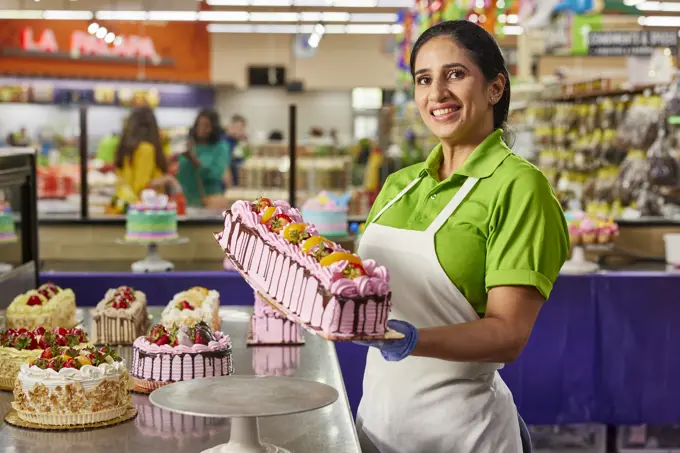 Female baker showing off finished layer cake after putting on the finishing touches of fresh fruit in latin market bakery.