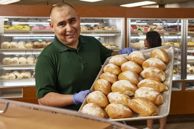 Portrait of Grocery store baker, smiling to camera refilling bin with tray of fresh baked Bolillos, a type of savory bread made in Mexico and Central America.