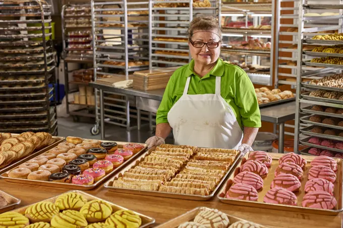 Portrait of female baker with apron and hair net in commercial kitchen of grocery store with large selection of freshly baked sweet breads surrounding her 