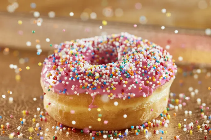 Close up detail view of donut with pink icing with decorative rainbow sprinkles falling into place 