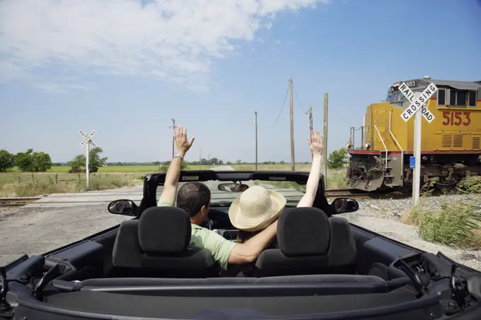 Middle aged baby boomer couple driving down country road in convertible with top down, Stopped at a railroad crossing waving to passing train 