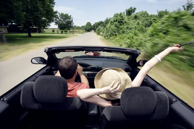 Middle aged baby boomer couple driving down country road in convertible with top down, woman with hand raised in the air. viewed from behind 