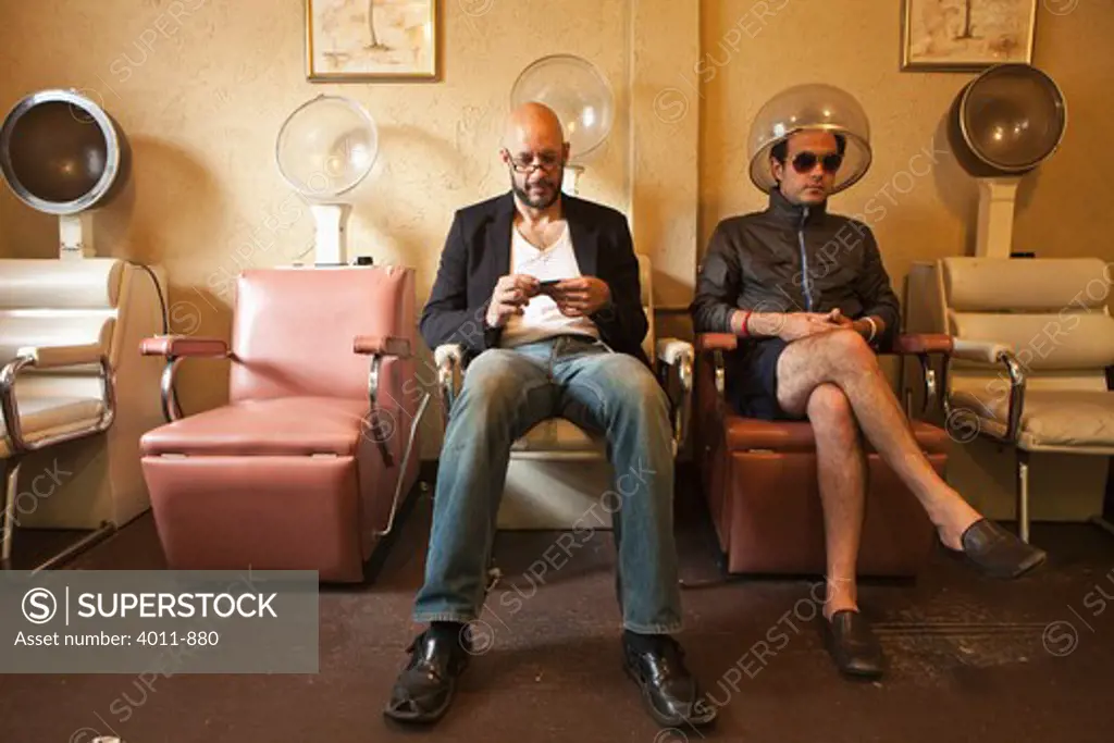 Two men sitting in old salon hair drying chairs