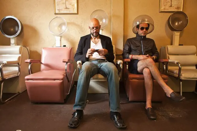 Two men sitting in old salon hair drying chairs