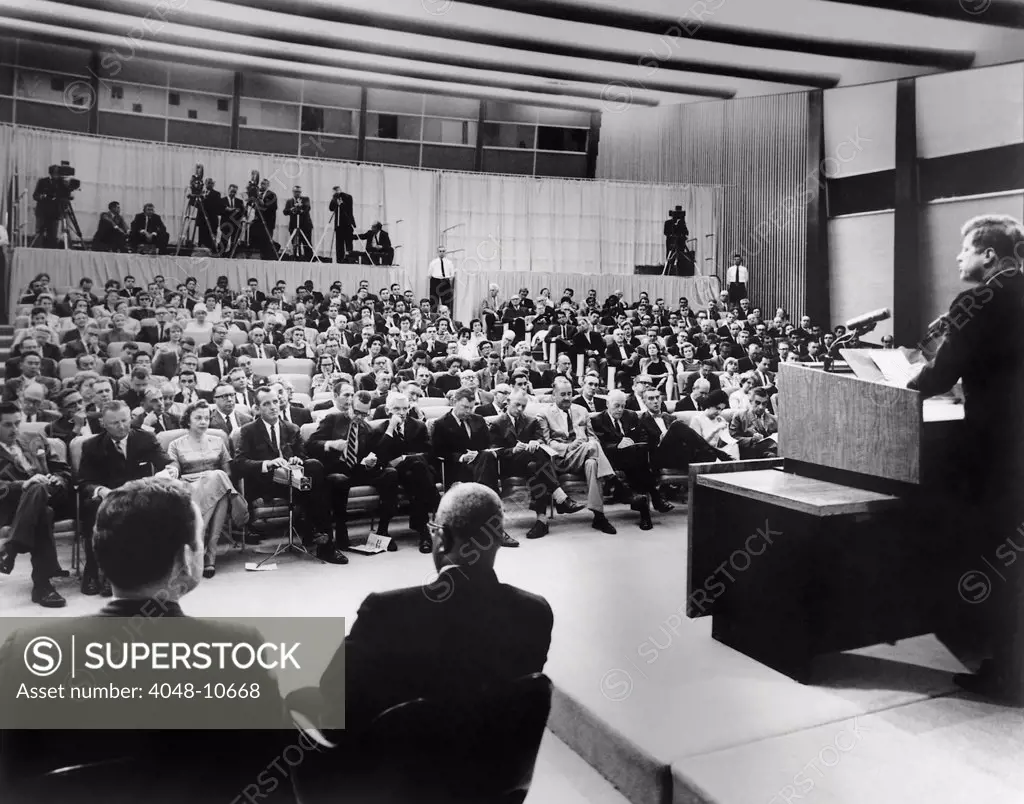 President John Kennedy speaks to reporters during a press conference. Press Secretary Pierre Salinger and Associate Press Secretary Andrew Hatcher sit near the podium at State Department Auditorium, Washington, DC. 1961-63.