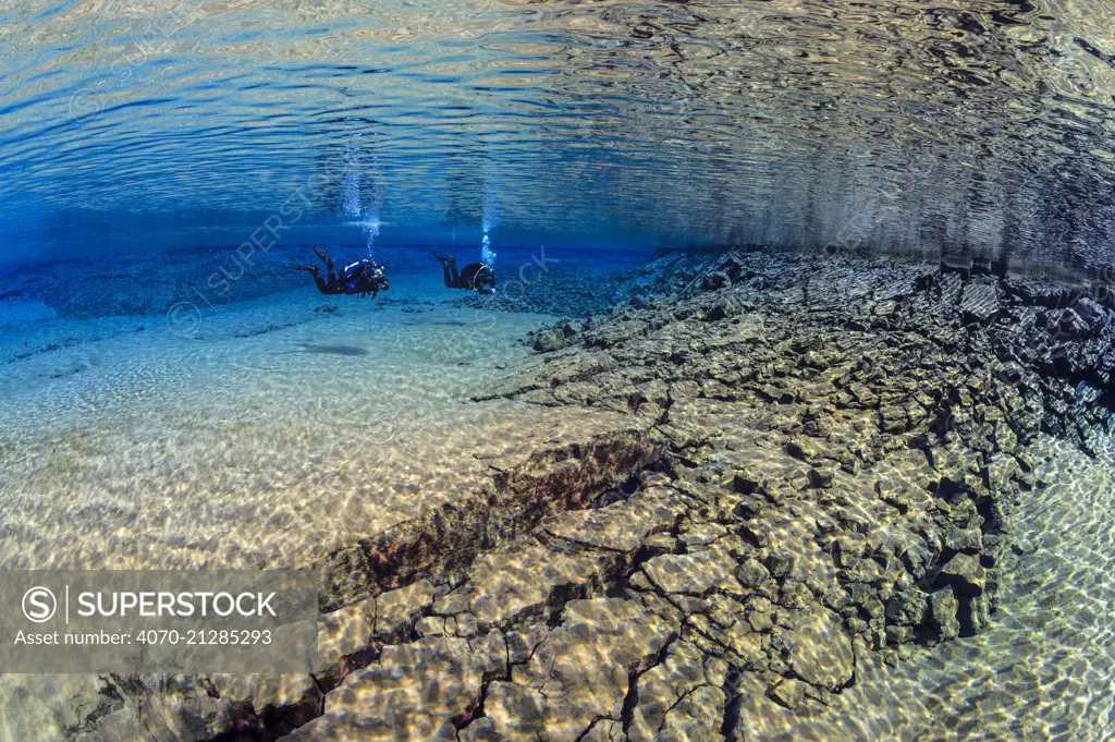 Pair of divers swimming in the blue lagoon at Silfraa, with fractured rocks in the foreground. Thingvellir National Park, Iceland. May 2011.   No model release available.