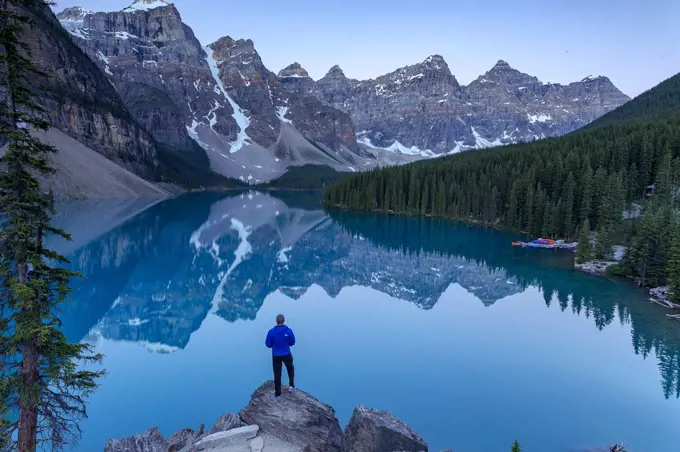 Person standing on rocks by looking over lake in the Rocky Mountains, with surrounding forest and kayaks stored, Alberta, Canada. July.