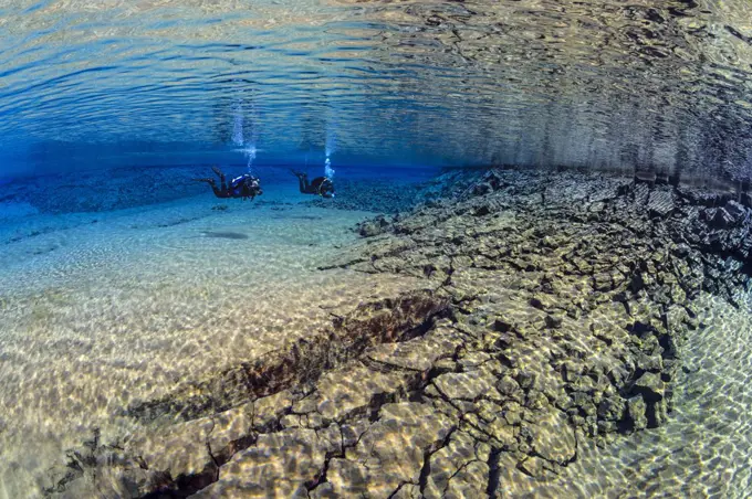 Pair of divers swimming in the blue lagoon at Silfraa, with fractured rocks in the foreground. Thingvellir National Park, Iceland. May 2011.   No model release available.