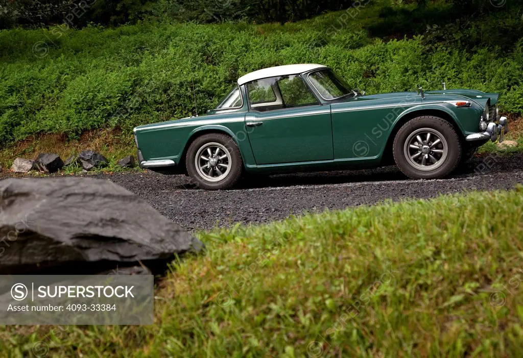 Profile view of a green 1967 Triumph TR4A IRS with a surrey top hardtop, parked in a lush natural location.