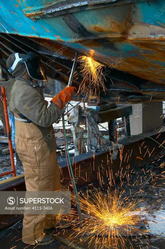 Worker in a shipyard working on the hull of a trawler with an acetelene torch.