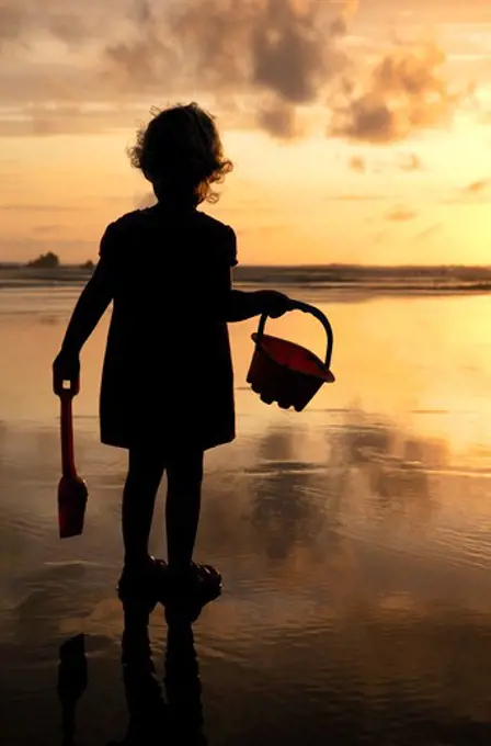 Little girl with bucket and spade silhouetted against setting sun on Summerleaze beach, Bude, North Cornwall, UK