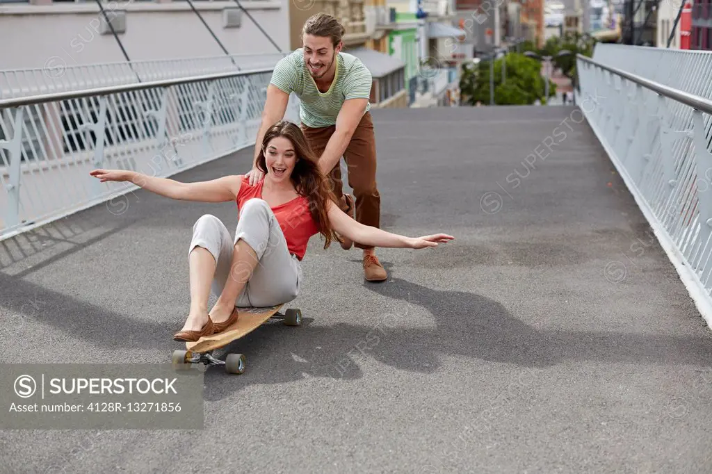 MODEL RELEASED. Young woman sitting on skateboard with man pushing.